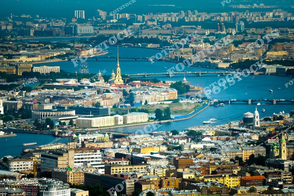 Вид на Санкт-Петербург с воздуха. В центре изображения расположена Петропавловская крепость на острове, окружённая реками и каналами. На переднем плане видны разноцветные здания, а на заднем - современные высотки и зелёные пространства. Городские мосты соединяют различные части города. Небо светлое, видно много зелени.